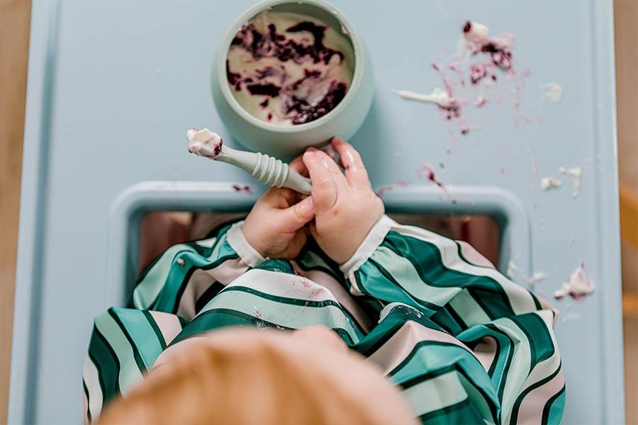 Baby etaing berries and yoghurt as a 'first meal' in highchair using Sticky Spoon and Suckie Scoop Bowl wearing Smockie Bib by Starting Solids Australia.