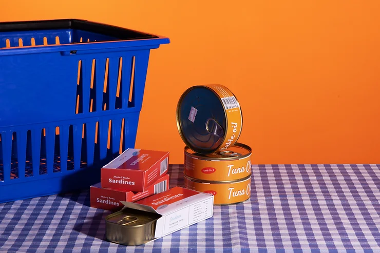 Cans of tuna and sardines stacked next to a blue shopping basket on a blue and white gingham tablecloth, in front of an orange wall.