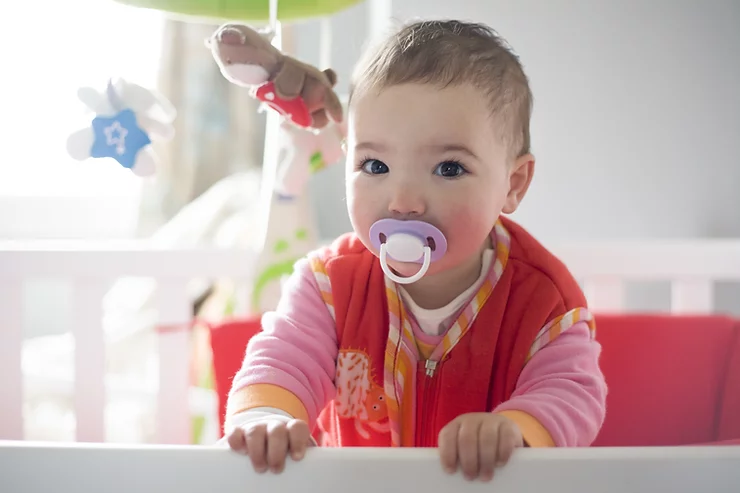 Baby with dummy wearing a red, pink and orange romper, standing up in a cot holding onto the edge.