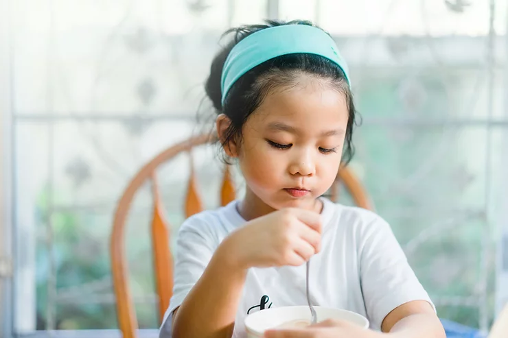 Young asian girl sitting at a table eating solid food from a bowl with a spoon.