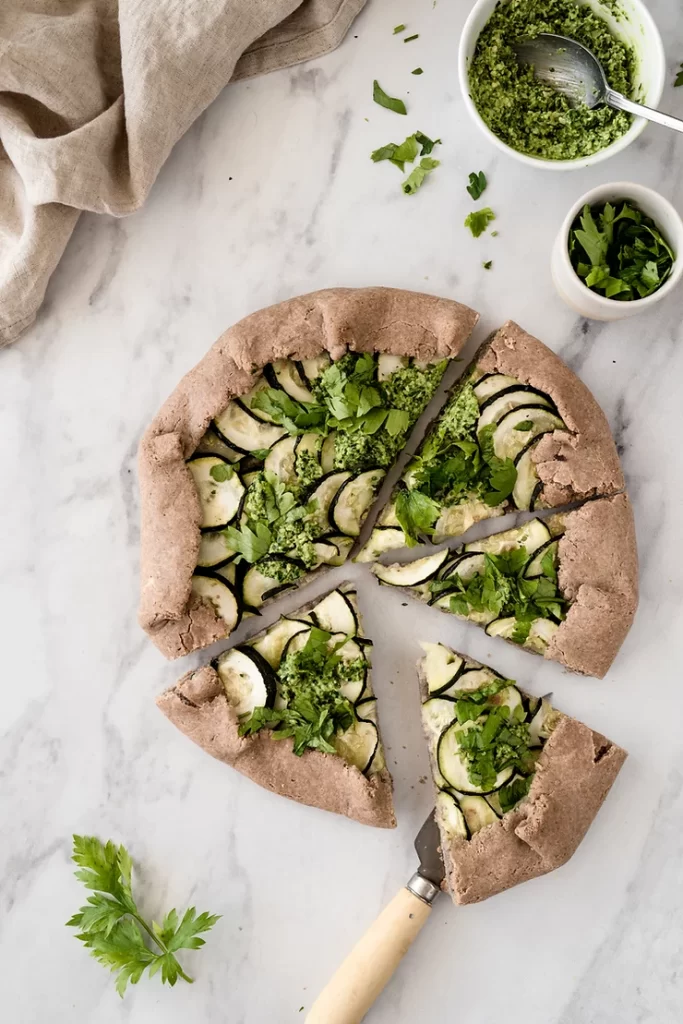 Pizza with green topping on marble bench with accompaniments.