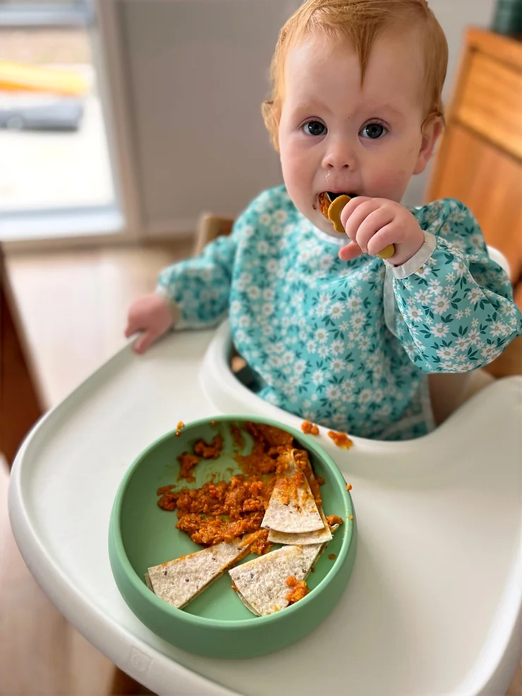 Baby girl eating red lentil mash in highchair wearing Starting Solids Australia Smockie Bib and using the Suckie Scoop Plate and Toddler Silvies.