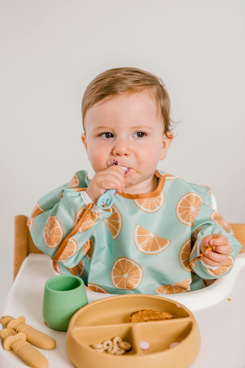 Baby boy in Tripp Trapp highchair eating solid food with Starting Solids Australia's silicone spoons, plates and cups whilst wearing a Smockie Bib in Oranges print.