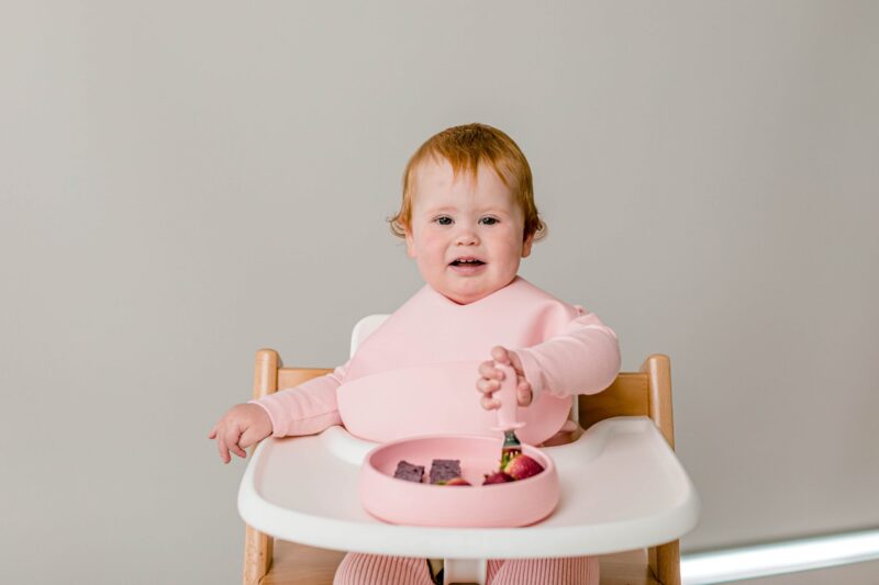 Baby girl sitting in Tripp Trapp highchair wearing Starting Solids Australia silicone Silly Bib and eating strawberries with Toddler Silvies out of a Suckie Scoop Plate.