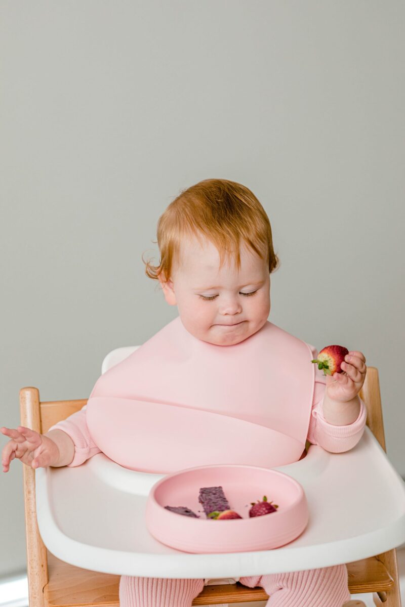 Baby girl eating strawberries in Tripp Trapp highchair wearing and using Starting Solids Australia silicone bib and plate.