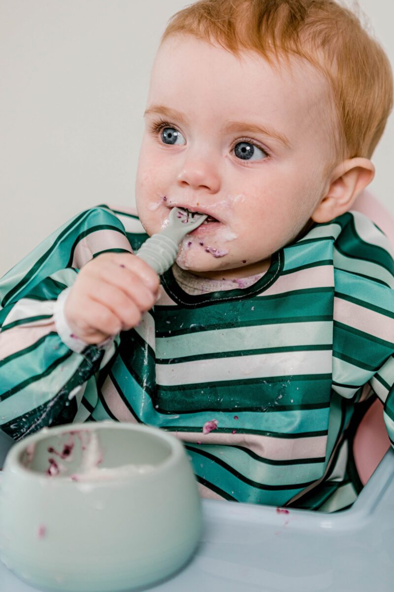 Baby boy eating solids with Starting Solids Australia's Stickie Spoon and Suckie Scoop Bowl, wearing a Smockie Bib in Aqua Lines print.