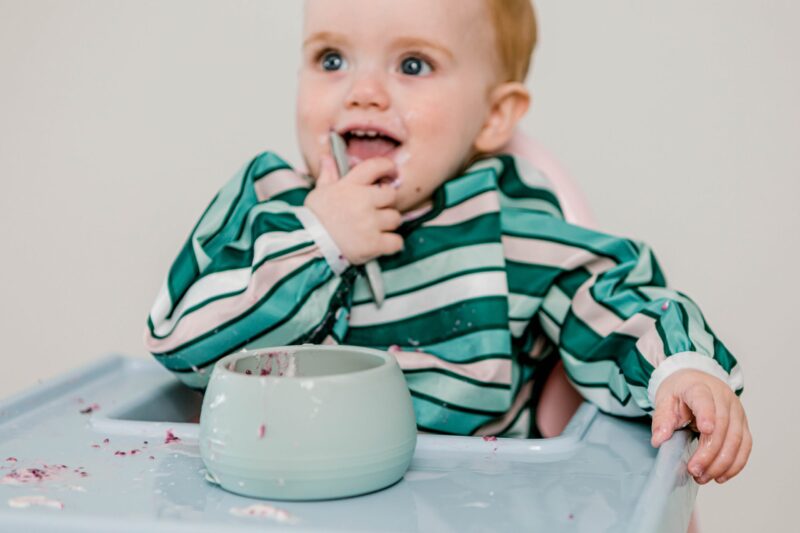 Baby boy sitting in a highchair eating solids with Starting Solids Australia's silicone Selfie Spoon and Suckie Scoop Bowl, wearing a Smockie Bib in Aqua Lines print.