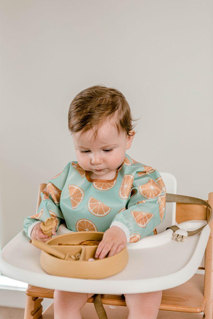 Baby boy picking solid food from Starting Solids Australia's Suckie Scoop Divided Plate, whilst holding a Selfie Spoon and wearing a Smockie Bib in Oranges print.