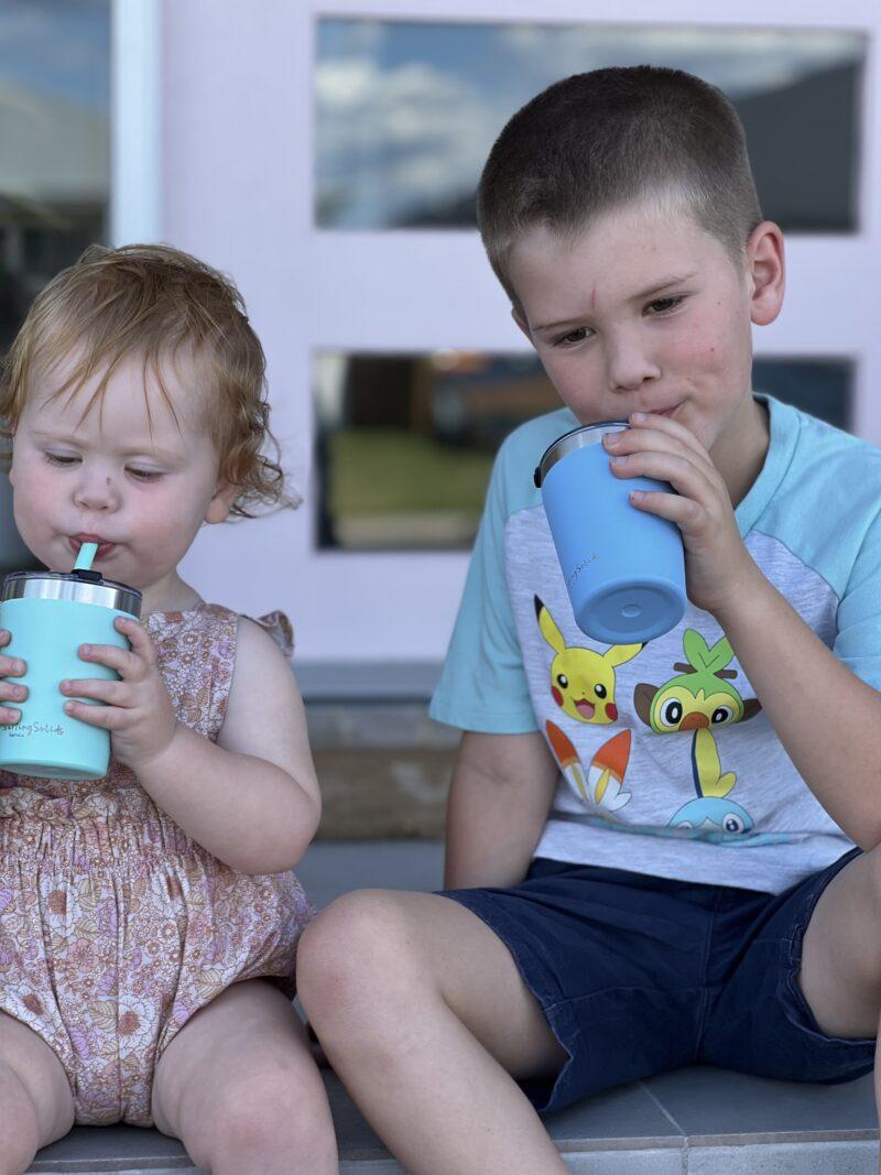 Toddler girl and boy drinking from a mint green and blue stainless steel insulated 'Big Kiddie Cup' with straw for kids by Starting Solids Australia.