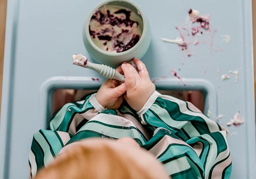 Baby etaing berries and yoghurt as a 'first meal' in highchair using Sticky Spoon and Suckie Scoop Bowl wearing Smockie Bib by Starting Solids Australia.