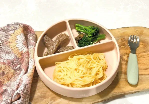 Beef and broccoli noodles in silicone Suckie Scoop Divided Plate with baby fork on wooden board with tea towel - Starting Solids Australia recipe.