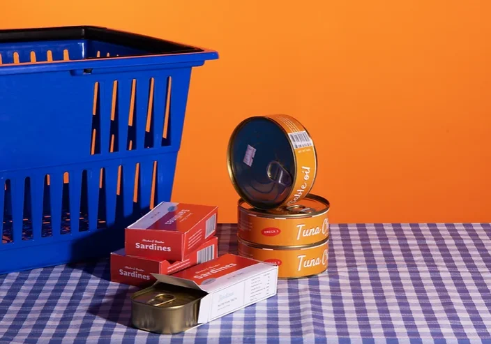 Cans of tuna and sardines stacked next to a blue shopping basket on a blue and white gingham tablecloth, in front of an orange wall.