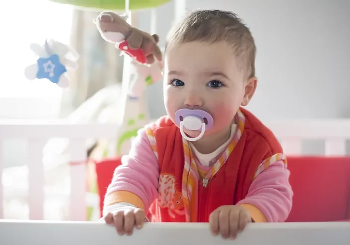 Baby with dummy wearing a red, pink and orange romper, standing up in a cot holding onto the edge.