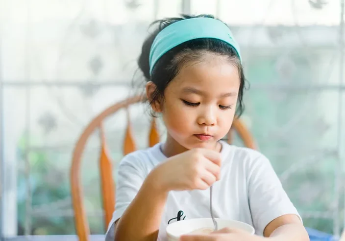 Young asian girl sitting at a table eating solid food from a bowl with a spoon.