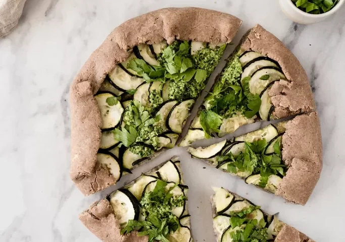 Pizza with green topping on marble bench with accompaniments.