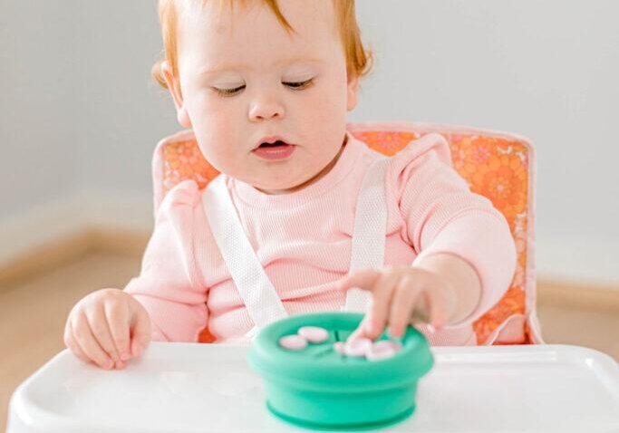 Baby girl sitting in Campie Chair by Starting Solids Australia in Retro Floral print, eating from a Snacie Cup.