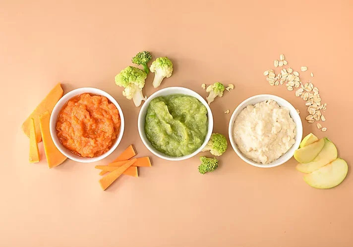 Pureed pumpkin, broccoli and oats with apple in three small white bowls on a peach background